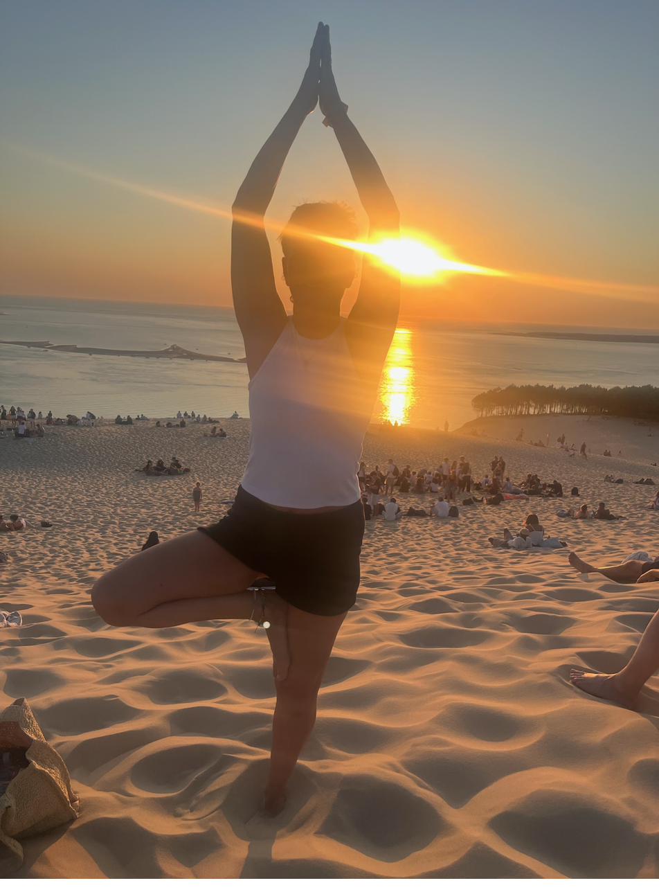 Vera steht in der Baumhaltung im Sand. Die tiefstehende Sonne leuchtet hinter ihr über dem Meer.