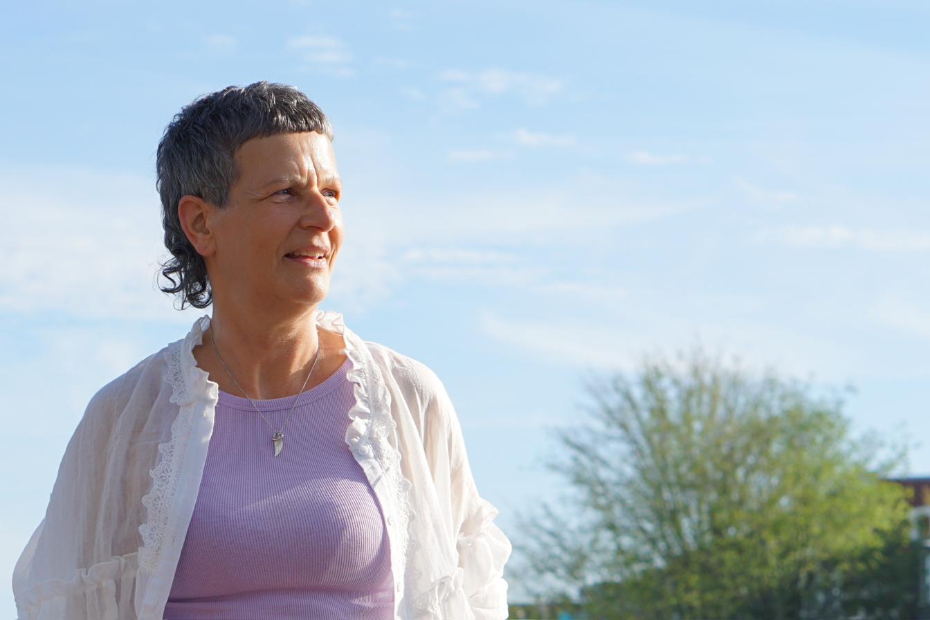 Vera lächelt in der Sonne, in einer hellen Bluse und einem lilafarbenen Oberteil.